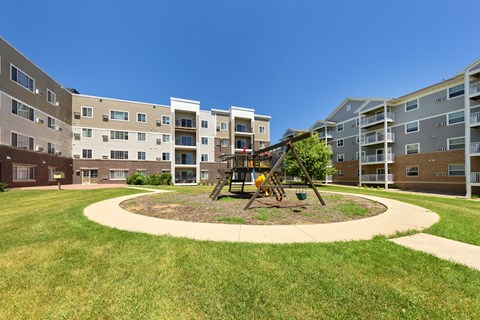 A playground with a swing set is in the middle of a grassy area in front of apartment buildings.
