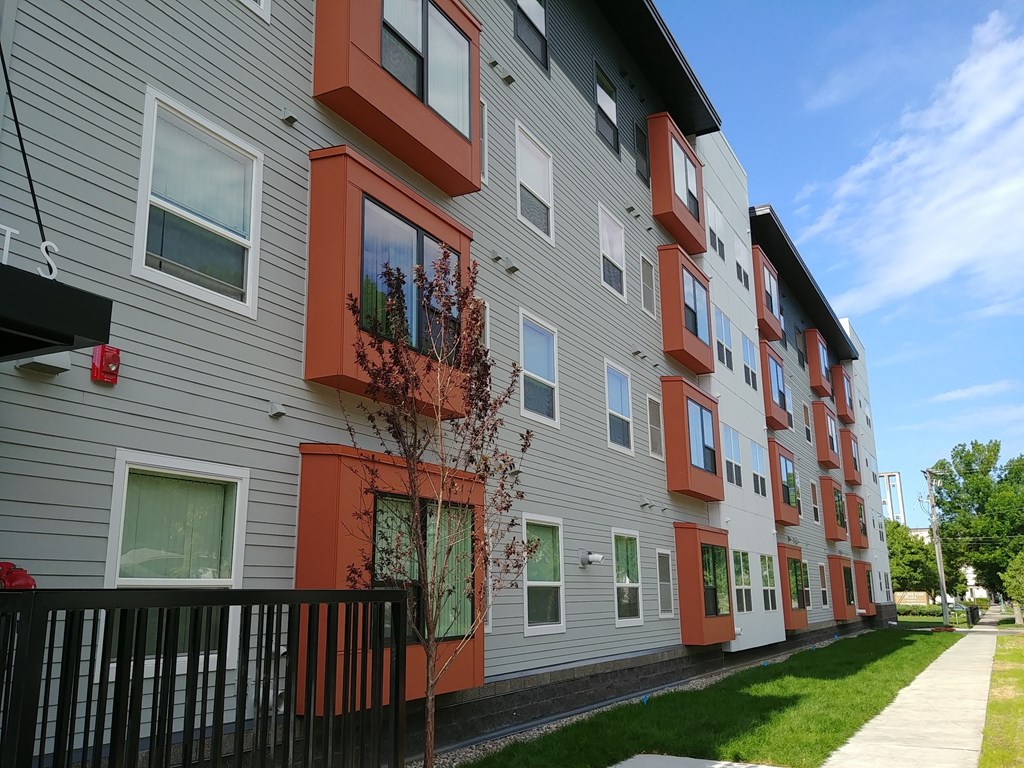 A tree with red leaves is in front of a grey building with orange trim.