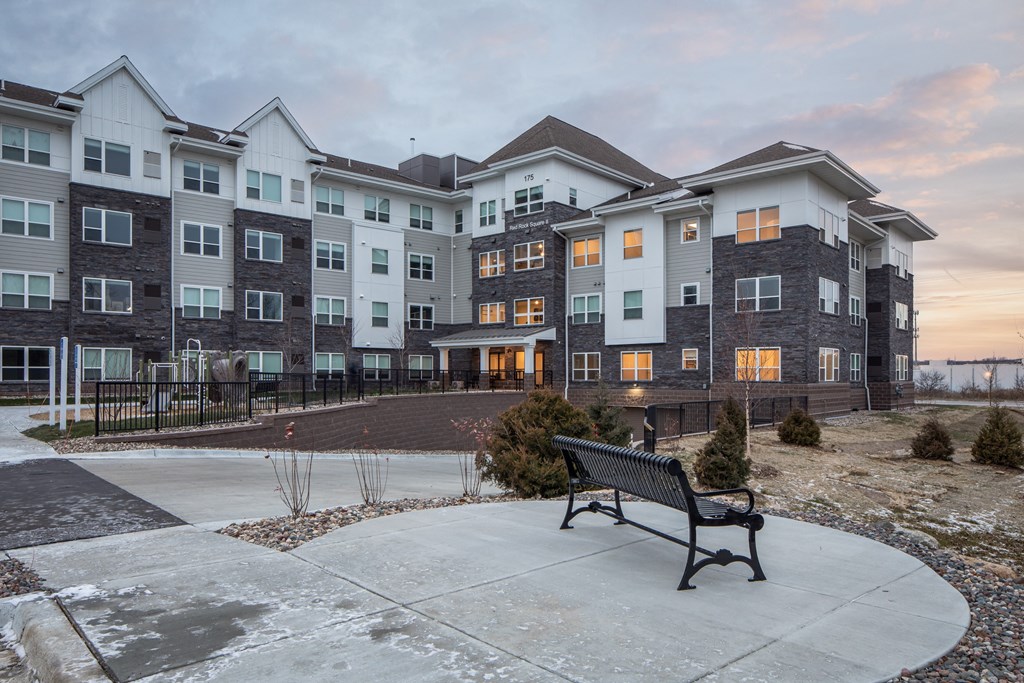 A bench sits in front of a building with a mix of white and grey balconies.