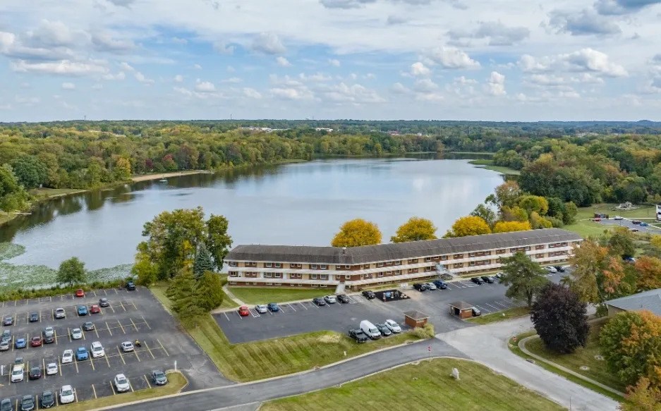 A large building is situated next to a parking lot with a lake in the background.