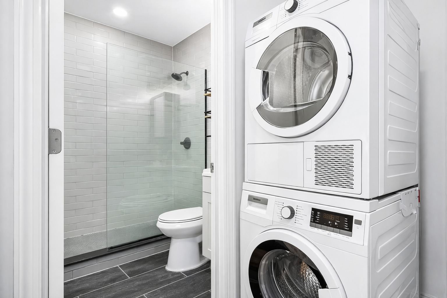 A white washing machine is in a bathroom with a toilet and glass shower door.