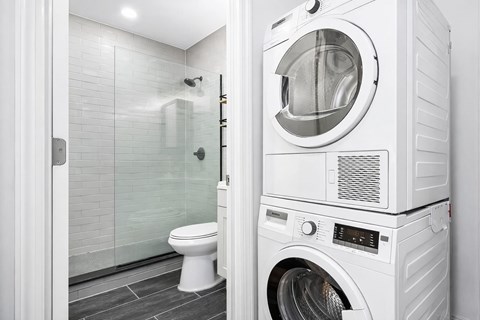A white washing machine is in a bathroom with a toilet and glass shower door.