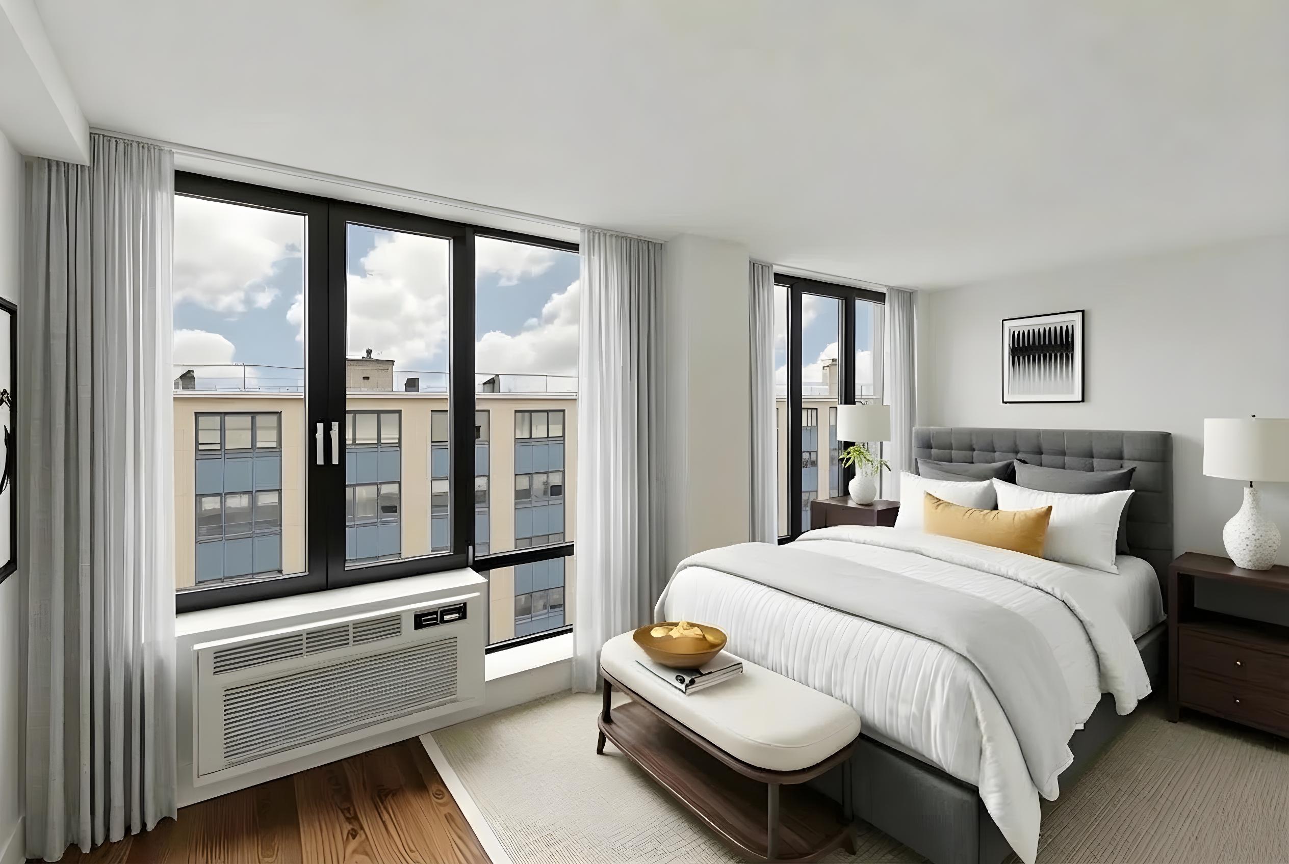 A bedroom with a large bed, a window with a view of a cityscape, and a nightstand with a lamp.
