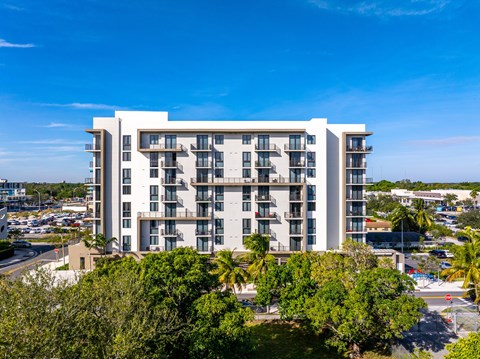 A large white building with balconies is surrounded by trees and a parking lot.