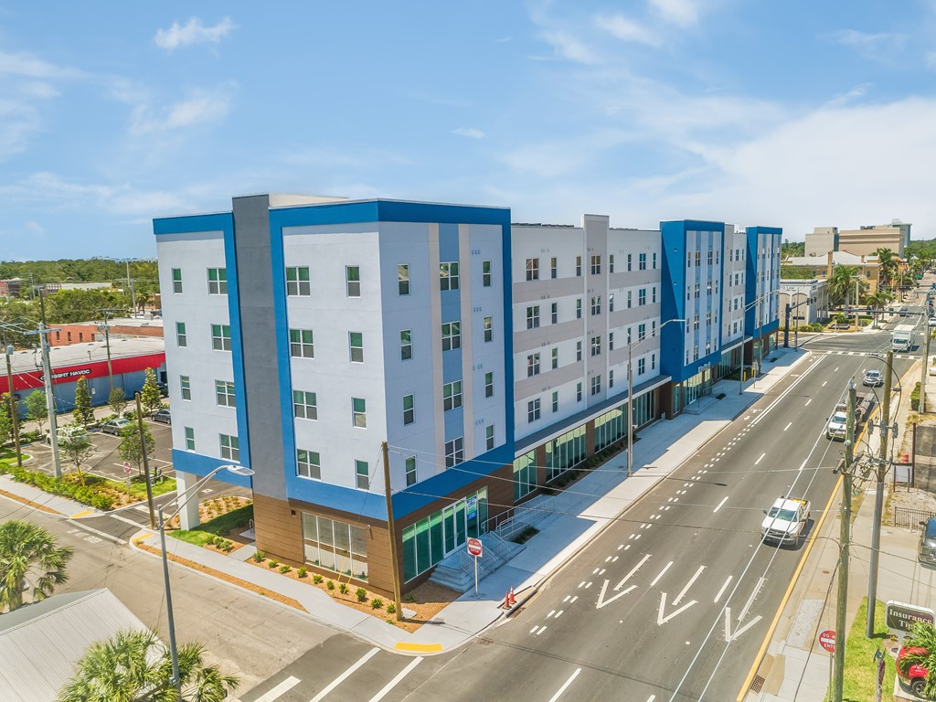 A modern multi-story building with blue and white exterior is situated on a street corner with cars parked along the curb.