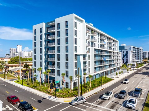 A modern white apartment building is situated on a street corner with cars parked and driving by.