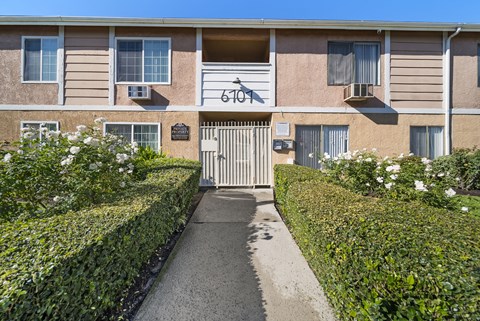 The image shows a building with the number 6107 on it, surrounded by a neatly trimmed hedge and a clear blue sky.