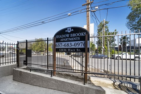 A sign for Shadowbrooks Apartments is displayed in front of a fence.