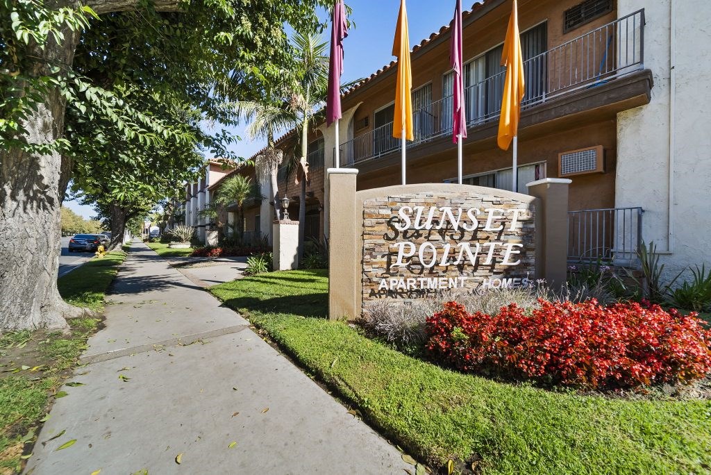 Sunset Polinte Apartment Homes sign with flags and red bushes.