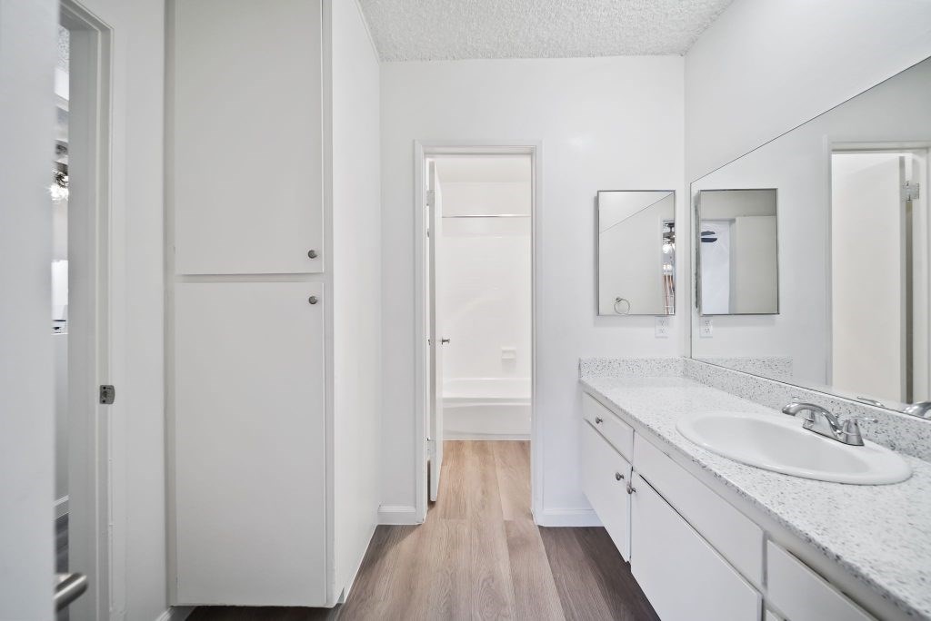 A white bathroom with a sink, mirror, and wooden floor.