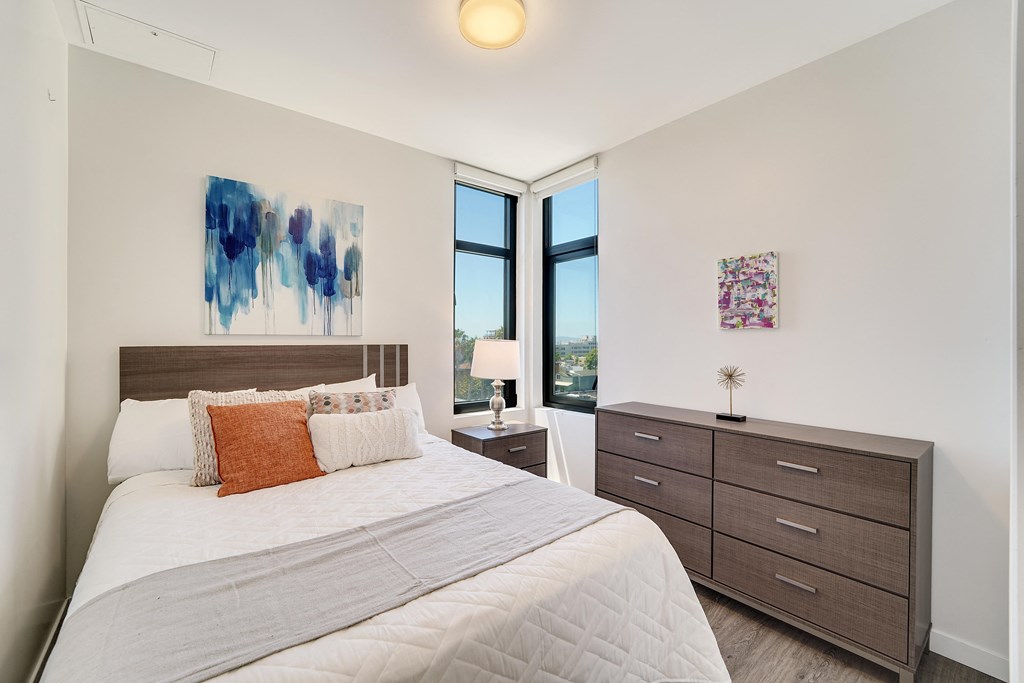 A bedroom with a large bed, a nightstand with a lamp, and two pieces of artwork on the wall.