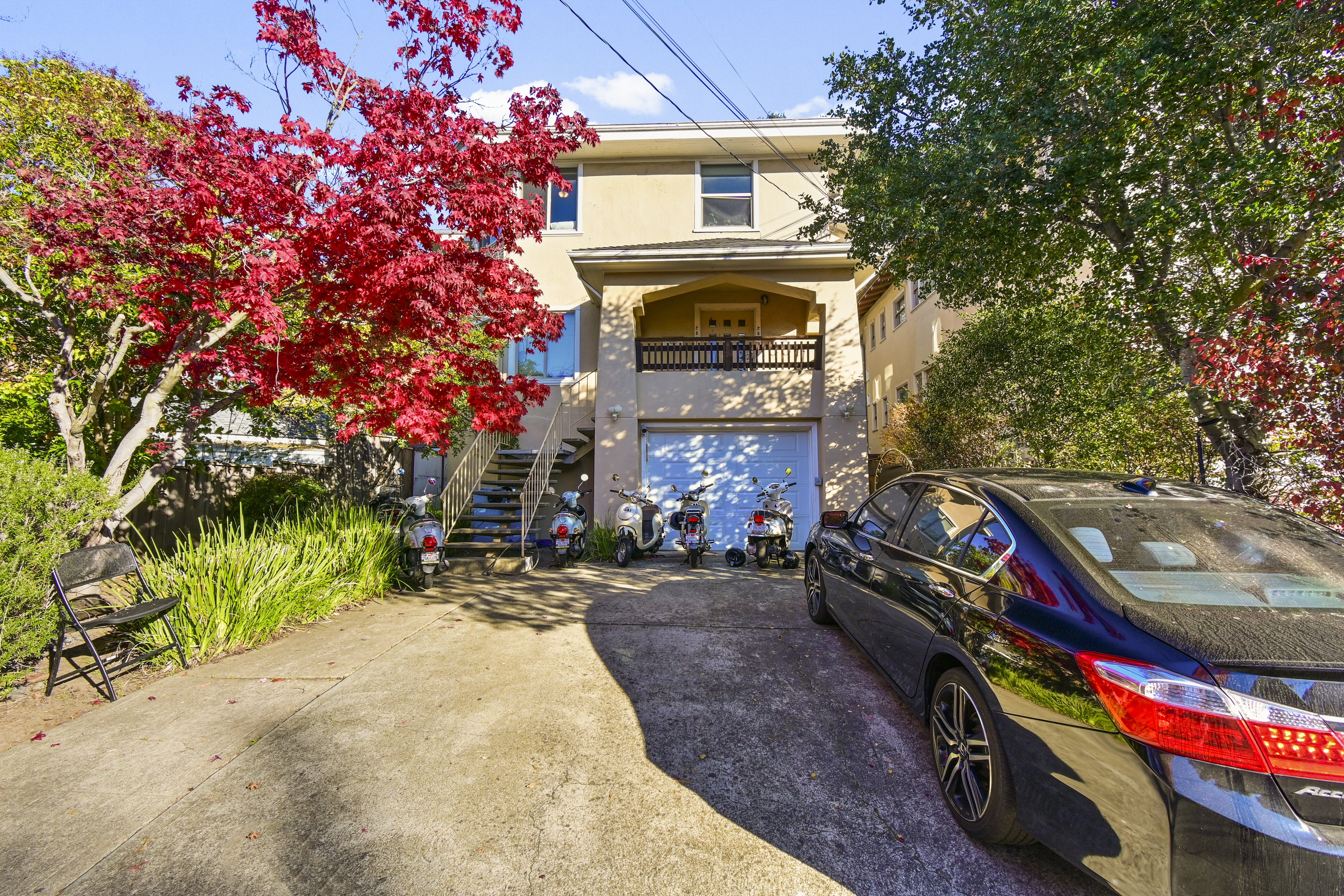 A car is parked in front of a house with a tree with red leaves.