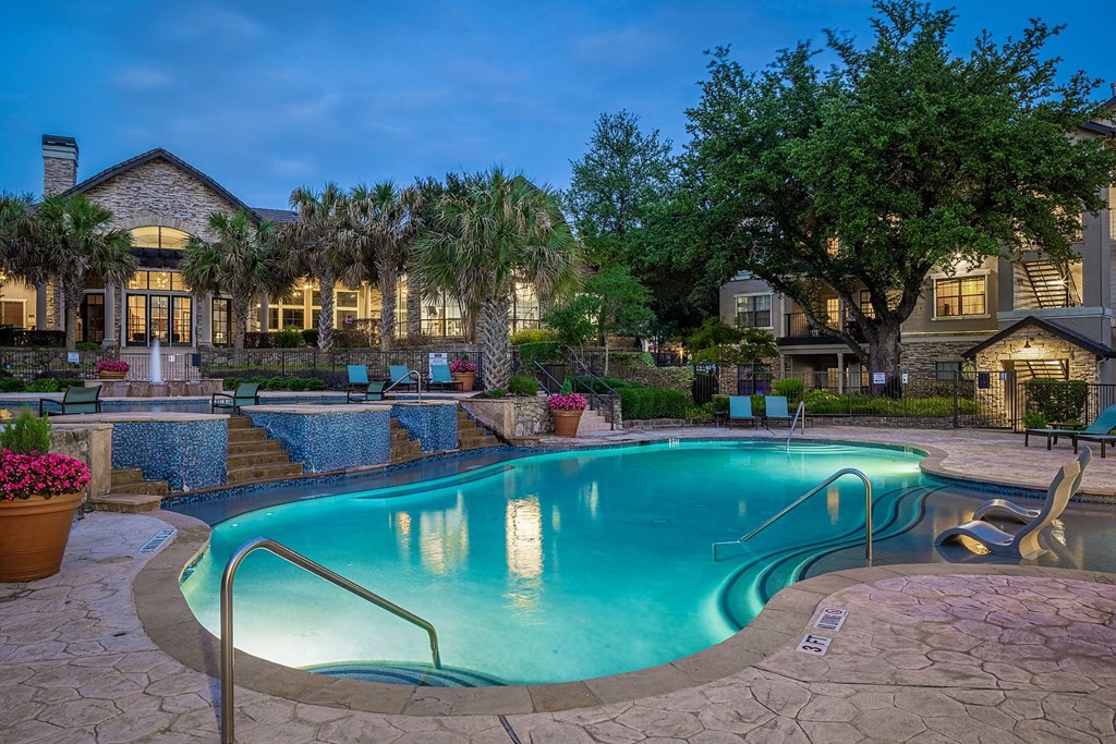 A swimming pool surrounded by a stone patio and a house with a lit up entrance.