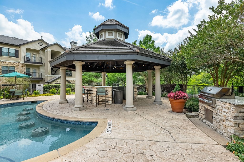 A gazebo is surrounded by a pool and a stone patio.