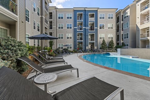 A pool surrounded by lounge chairs and umbrellas in front of apartment buildings.