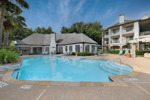 A large swimming pool in front of a building with a patio and a tree.