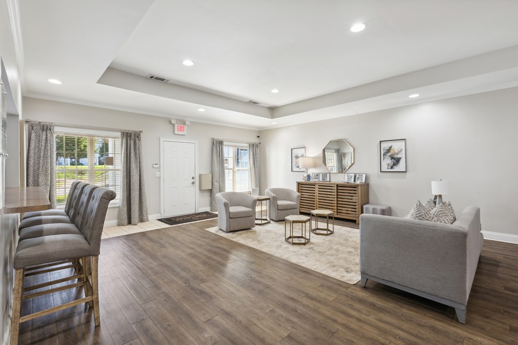 A well-lit, spacious living room with a grey sofa, wooden chairs, and a rug.