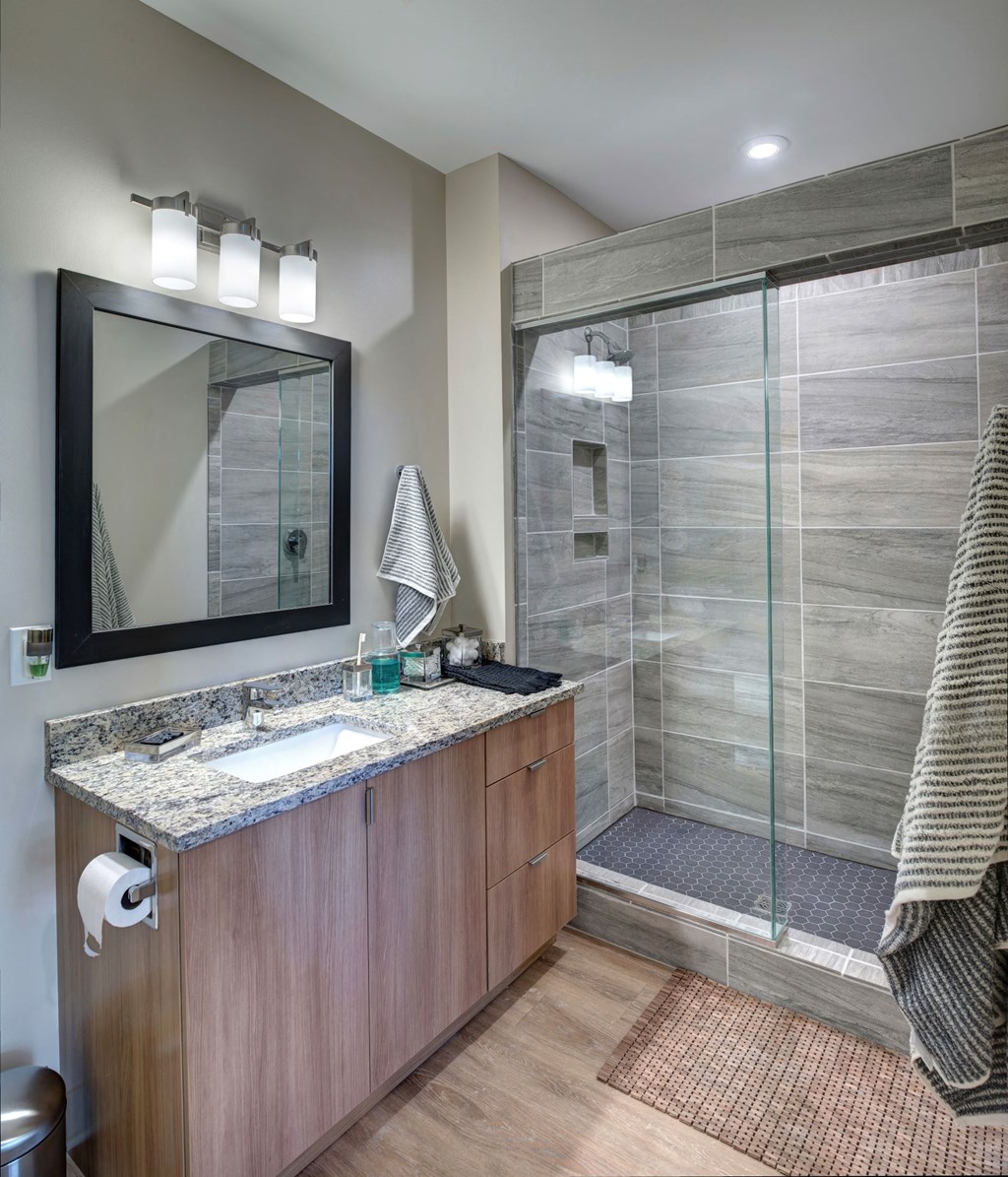 Apartments For Rent In Downtown Kalamazoo, MI - Large Bathroom With Grey Tile Shower And Glass Doors, Granite Countertops, Large Vanity Mirror, And Wood-Inspired Cabinets And Flooring