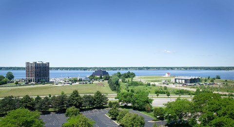 A large building sits on the left side of a body of water, with a parking lot and trees in the foreground.