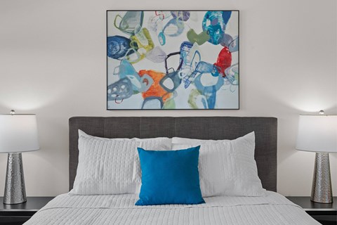 A bed with a white comforter and a blue pillow is in front of a painting.