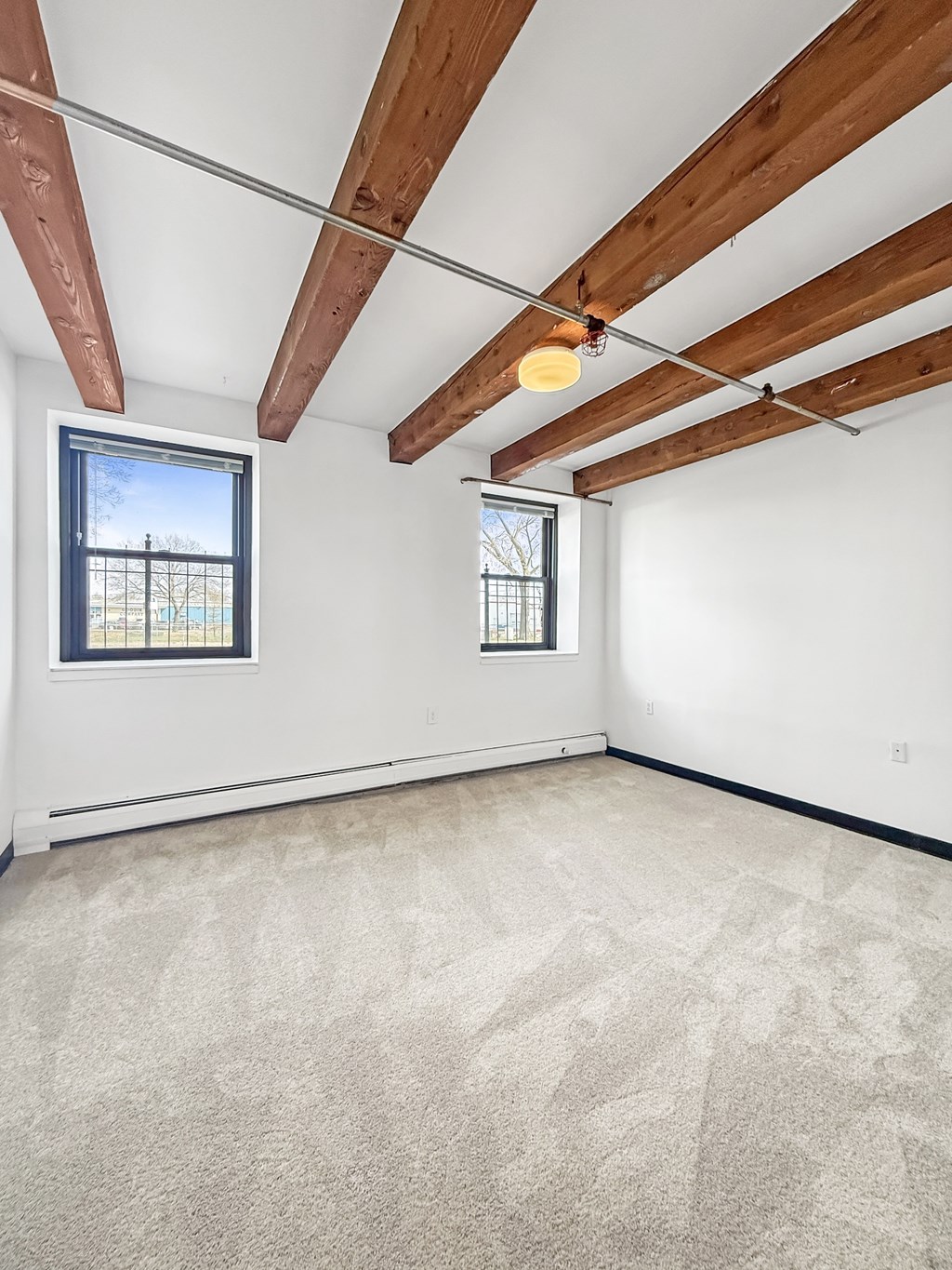 A room with a carpeted floor, wooden beams on the ceiling, and two windows.
