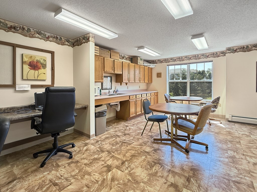A room with a desk, chairs, and a kitchen area.