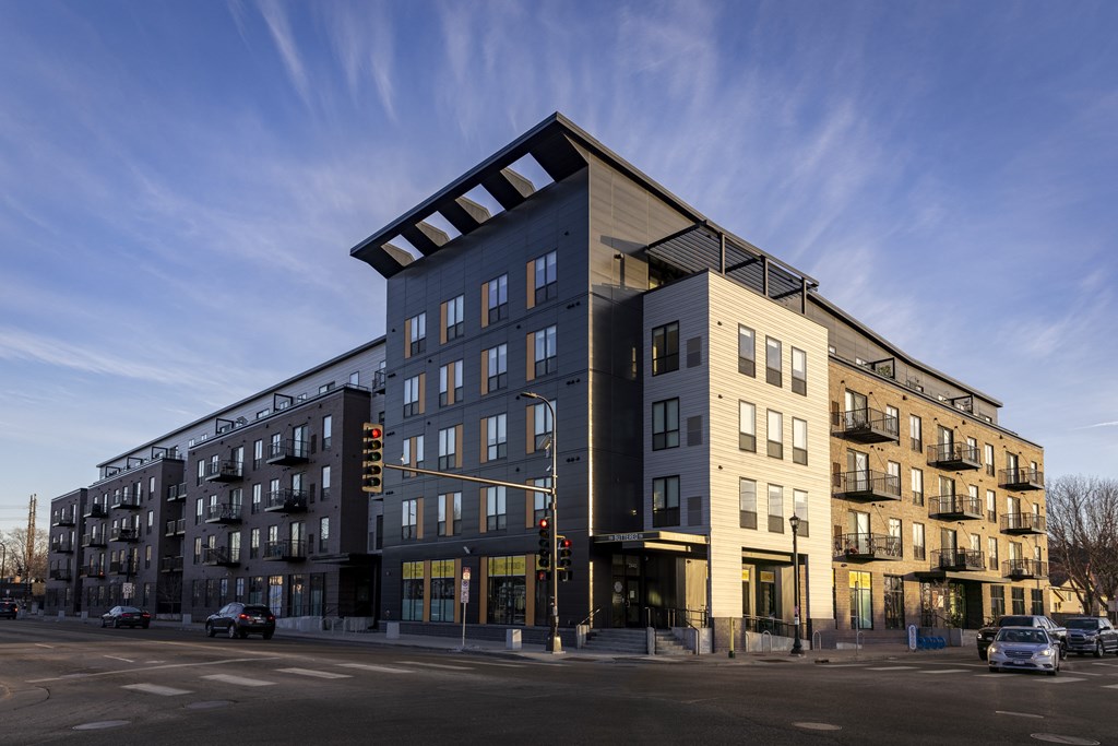 A modern apartment building with a mix of dark and light facades is situated on a street corner with cars parked along the curb.