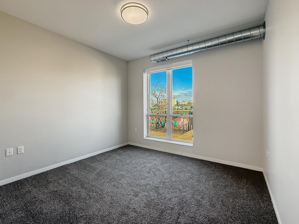 A room with a grey carpet and a window showing a view of a tree and a building.