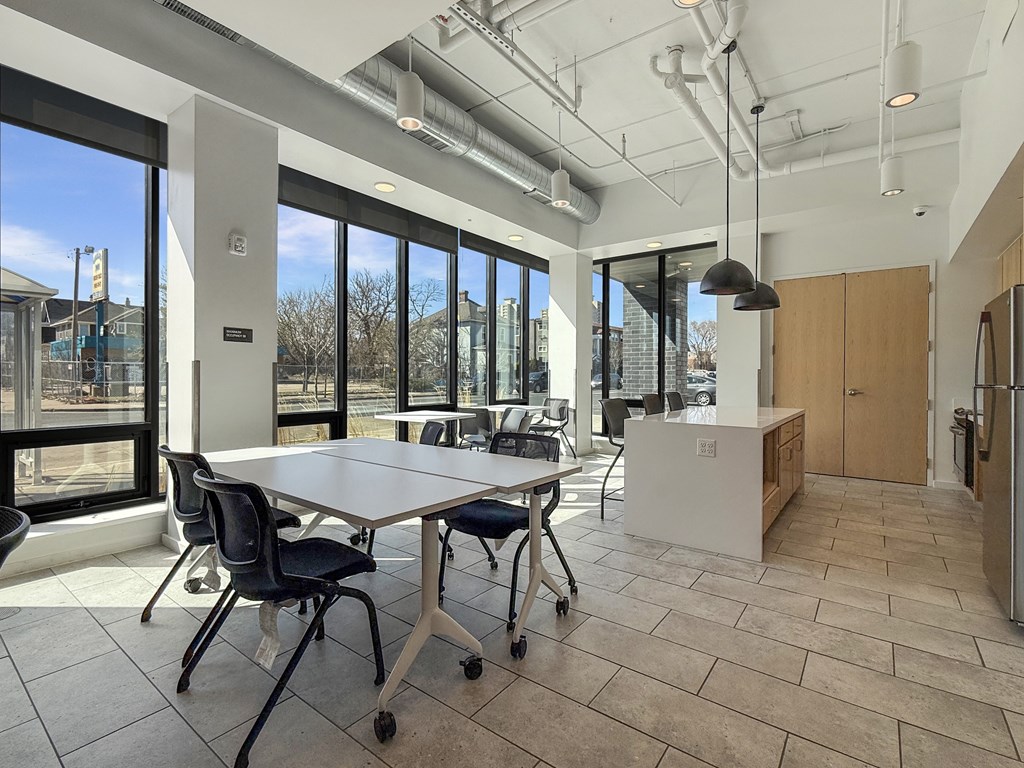 A conference room with a long table and chairs, a kitchenette, and a view of the outside through large windows.