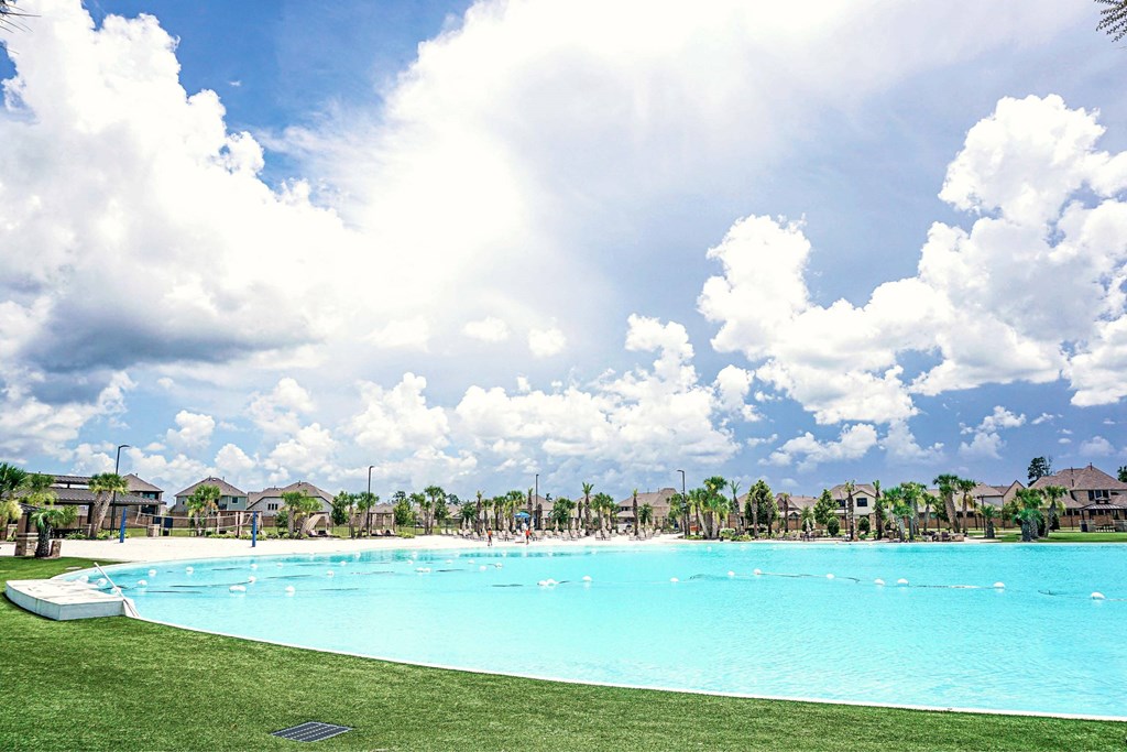 A large swimming pool surrounded by a grassy area and palm trees under a blue sky with clouds.