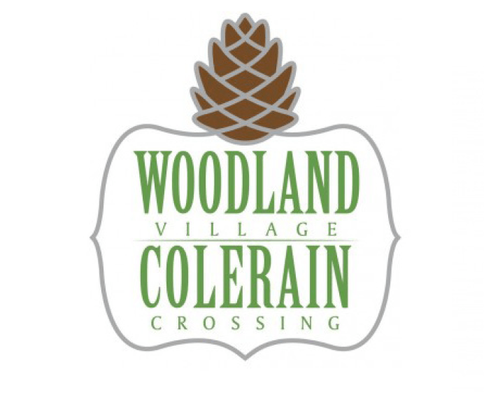 A logo with a pine cone on top and the words "Woodland Village Colerain Crossing" written below it.