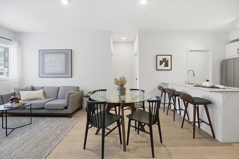 A modern dining room with a grey couch, a black table and chairs, and a white kitchen area.