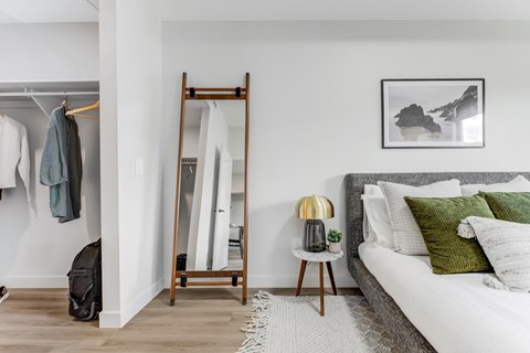 A bedroom with a bed, a clothes rack, a side table with a lamp, and a painting on the wall.