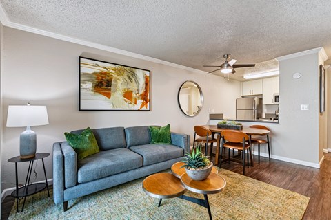 A living room with a grey couch, a painting on the wall, a round mirror, and a dining area in the background.