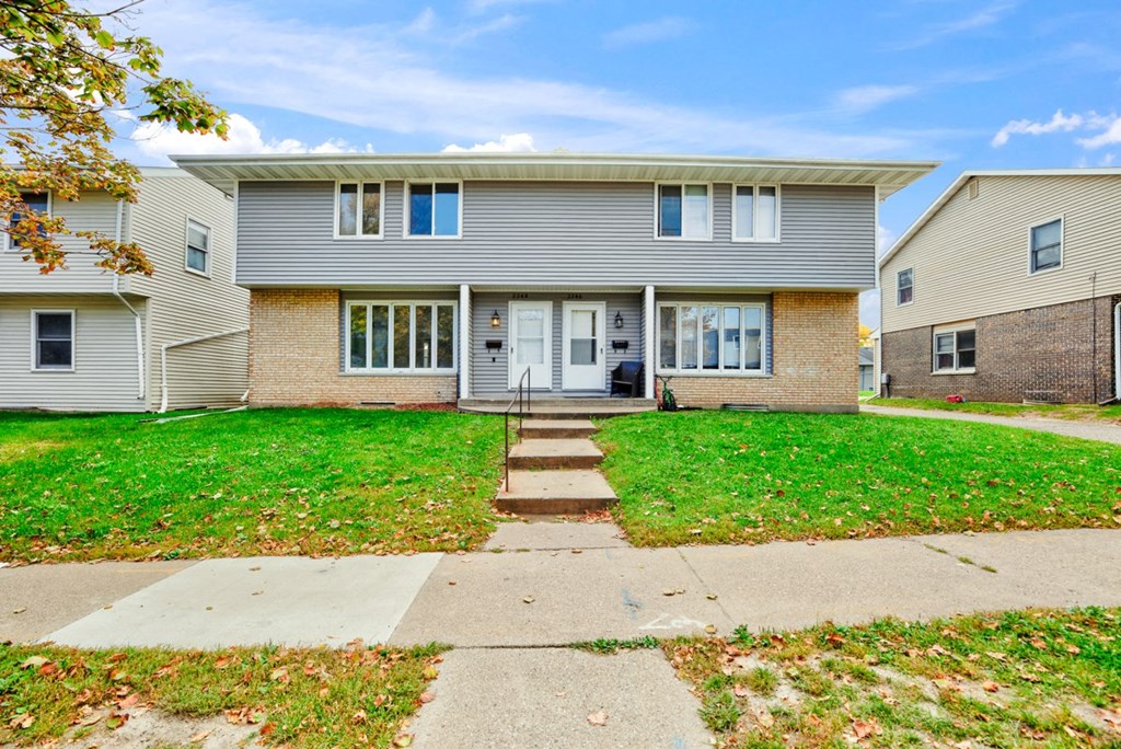 A house with a grey facade and a white door is surrounded by a green lawn.