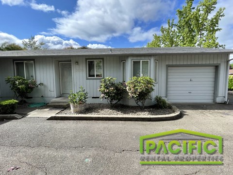 a small gray house with three bushes in front of it