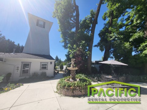A white house with a tower-like structure on the roof is surrounded by greenery and has a sign that reads "PACIFIC RENTAL PROPERTIES".