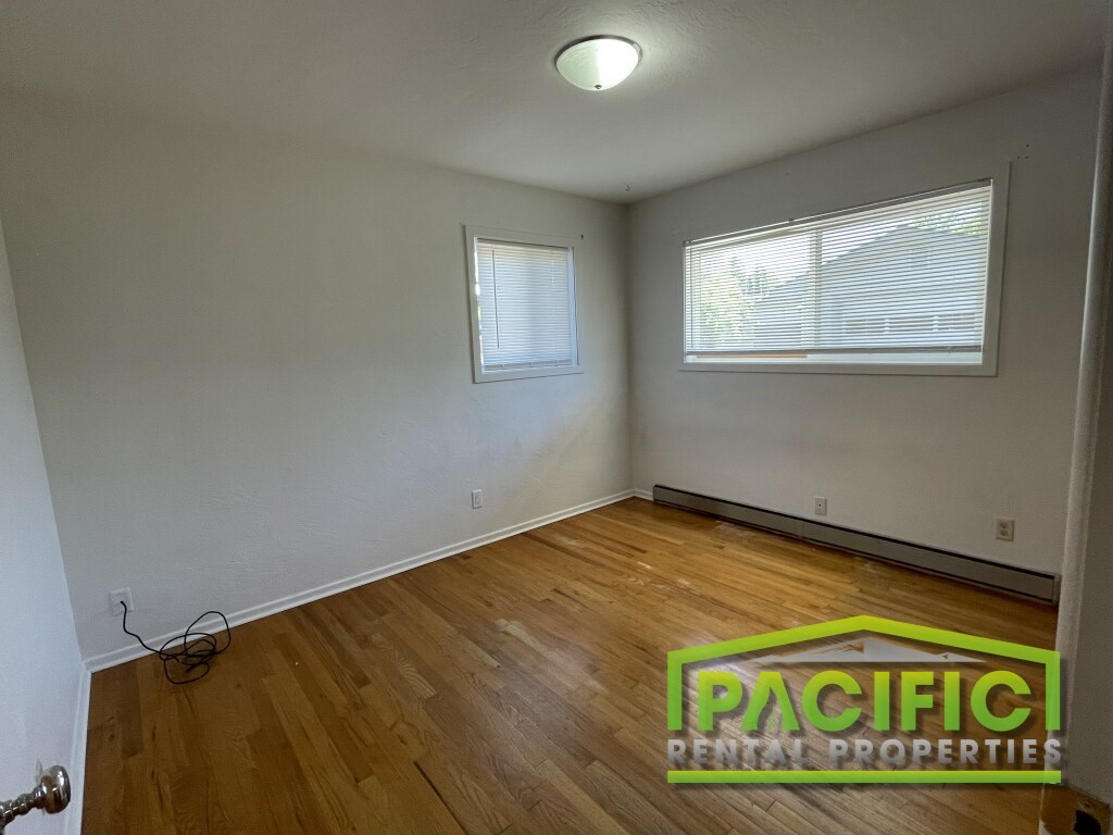 A room with wooden flooring and two windows, with a Pacific Rental Properties logo in the corner.