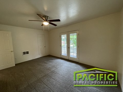A room with a carpet floor, a ceiling fan, and a window with a view of trees.