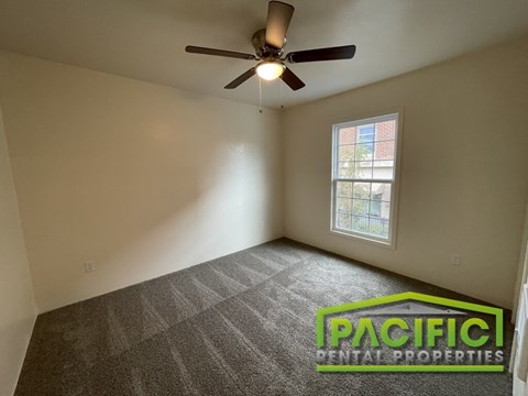 A room with a carpet floor, a ceiling fan, and a window with a view of a building outside.