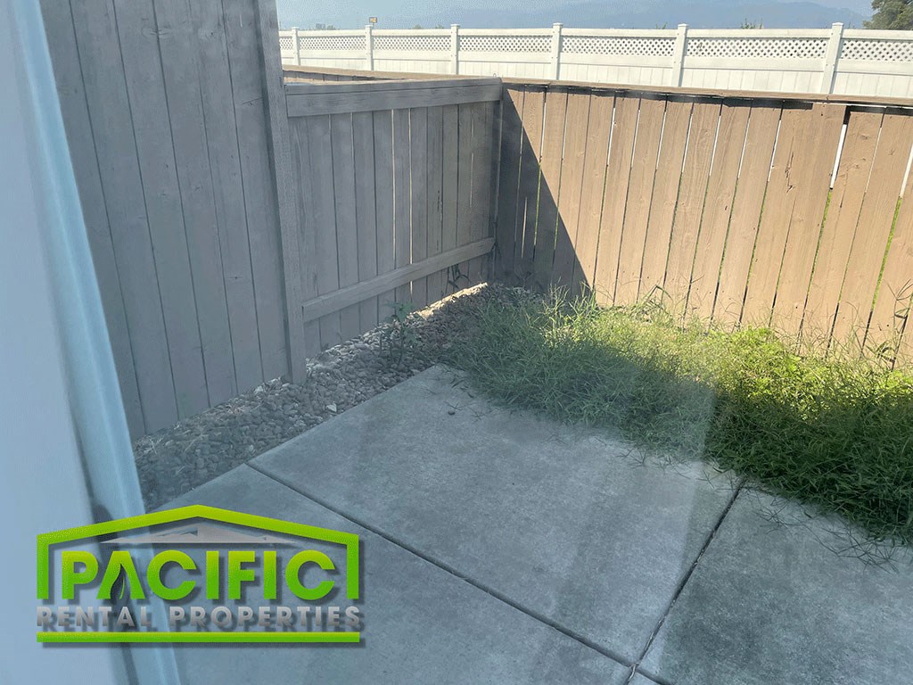 a corner of a backyard with a fence and a sidewalk