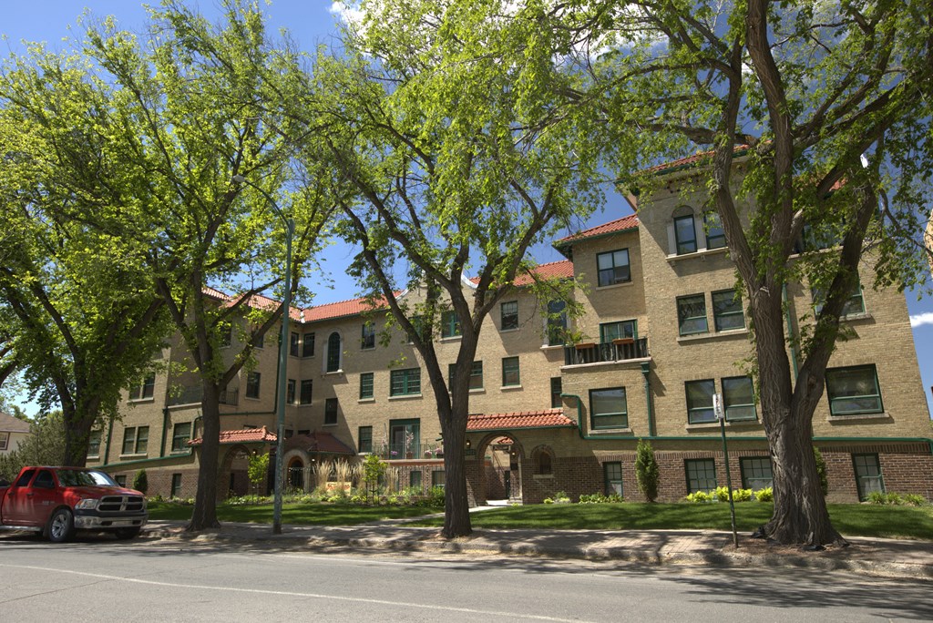 A red car is parked on the street in front of a building with a tree in front of it.