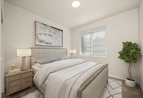 A bedroom with a large bed, a nightstand with a lamp, a plant, and a framed artwork on the wall.