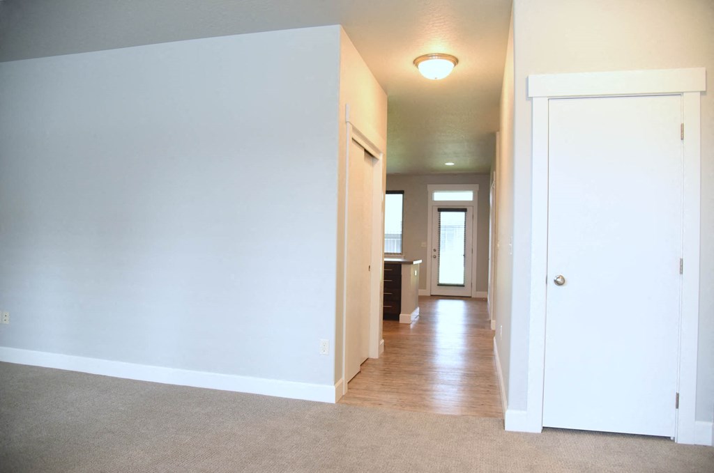 an empty living room with a hallway to a kitchen and a door to a house