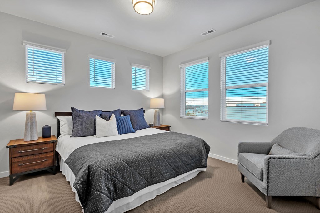 A bedroom with a large bed, a chair, and two windows with blinds.