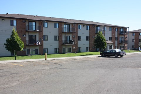 A black car is parked in a parking lot in front of apartment buildings.