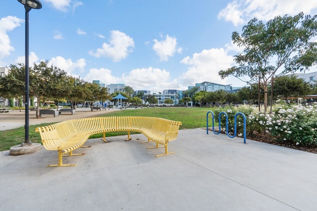 A yellow bench sits in a park with a blue pole behind it.