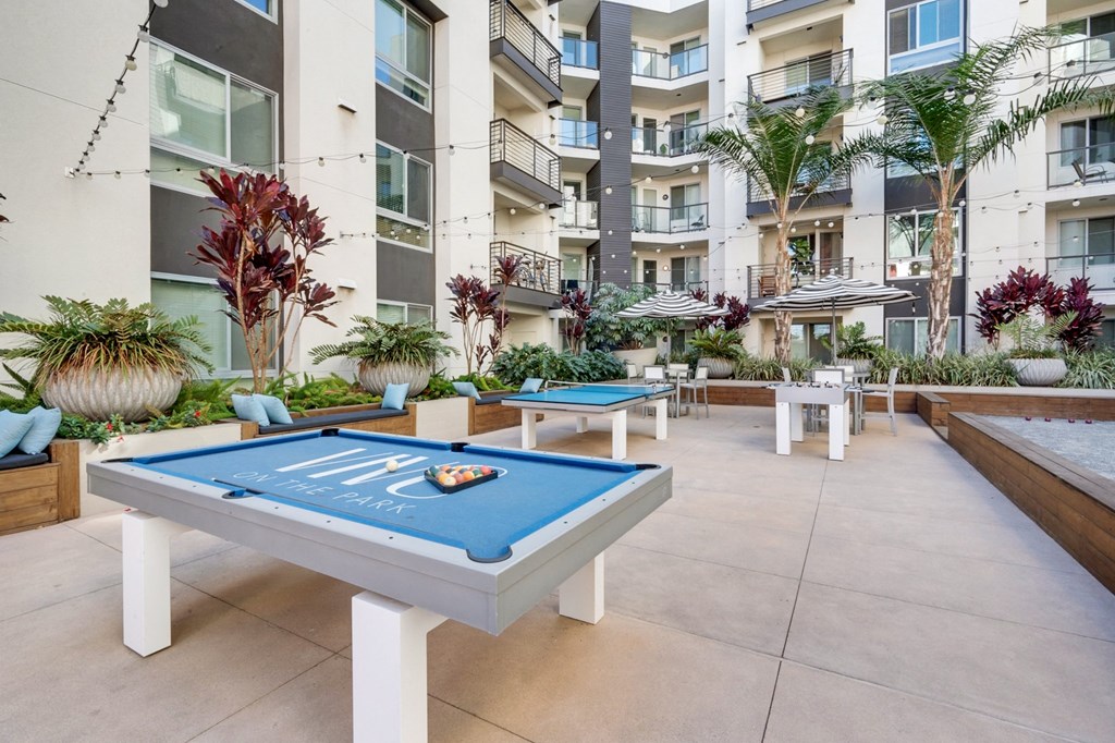 A pool table with a blue cloth and white legs is in the middle of a courtyard with plants and a building in the background.