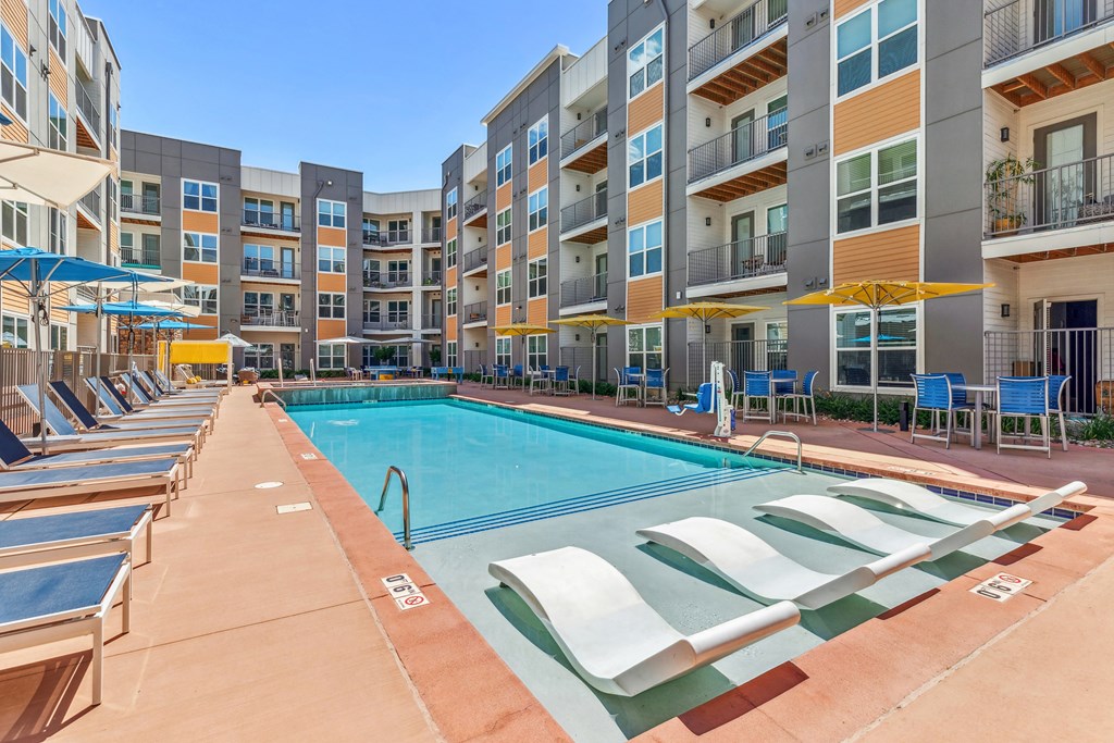 a swimming pool with chaise lounge chairs and umbrellas in front of an apartment building