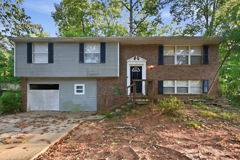 A two-story house with a garage is surrounded by trees and has a brown driveway.